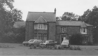 Black and white grainy image of a grand building with large windows. Old vehicles are parked outside next to a grass lawn.