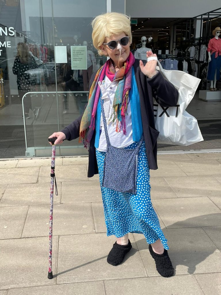 Susan stands outside a glass fronted shop with a shopping bag slung playfully over her shoulder. She is dressed in colourful clothes and is smiling at the camera. She is wearing sunglasses and using a walking aid.