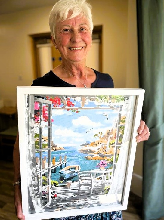 woman proudly displaying a framed painting of a scenic view.