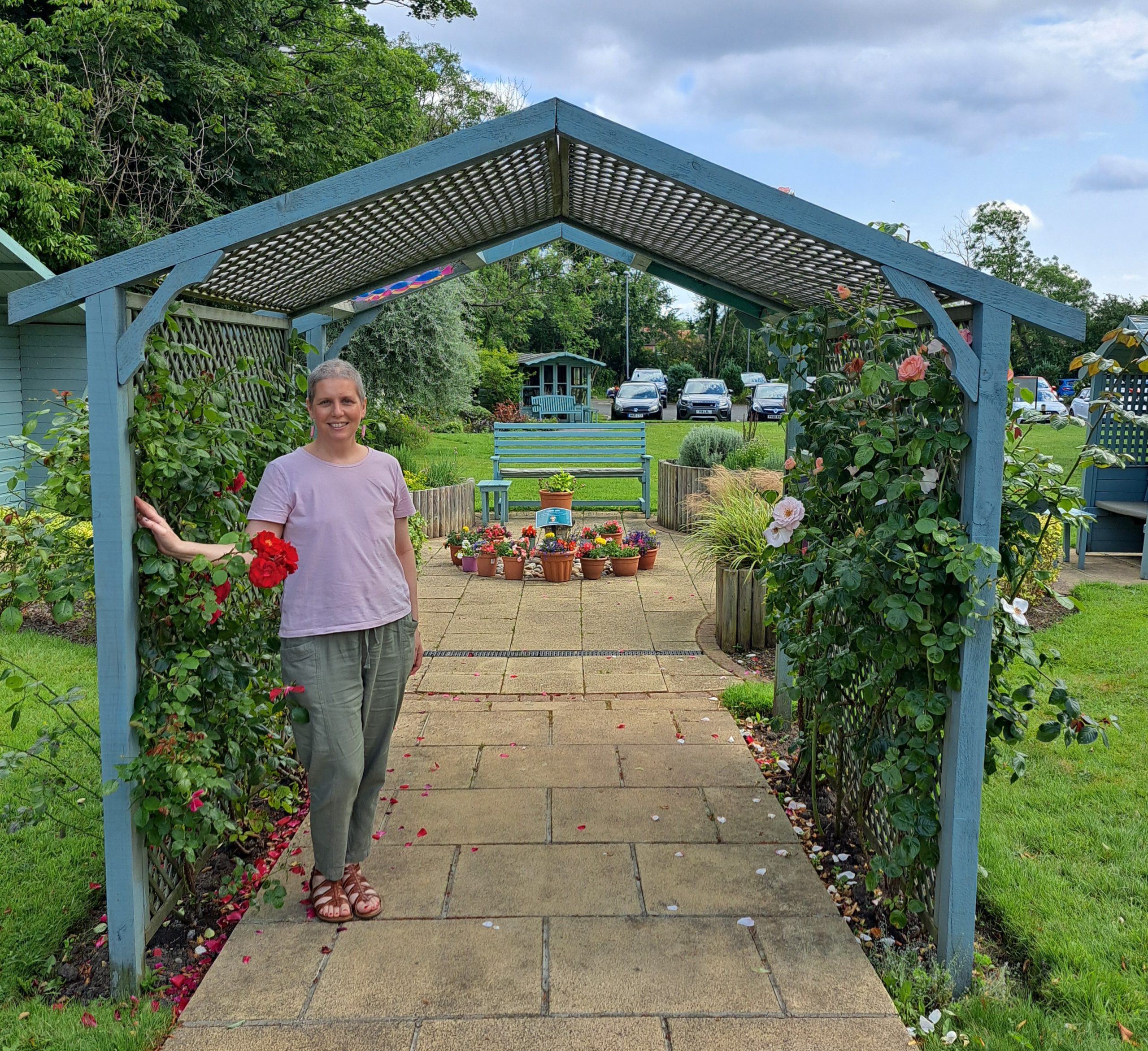 A woman smiles while standing in front of a picturesque gazebo, framed by vibrant flowers and greenery.