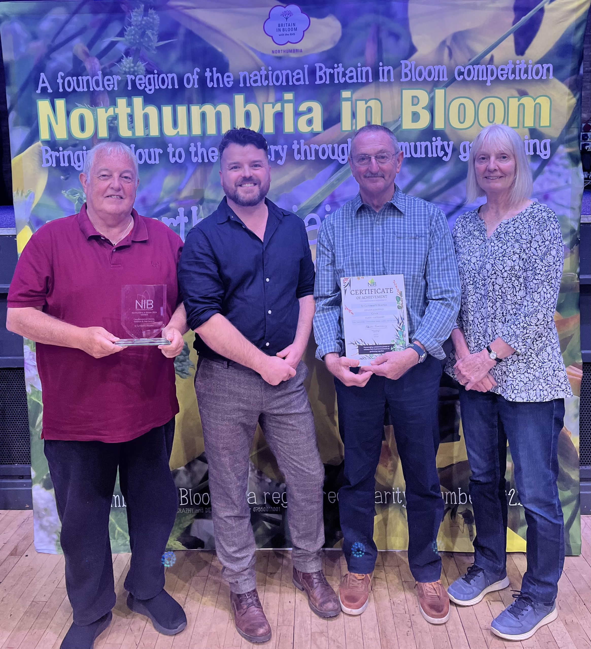 Four smiling individuals, three represent St Cuthbert's Hospice and are proudly holding an award for Northumberland in Bloom, celebrating their achievement together.