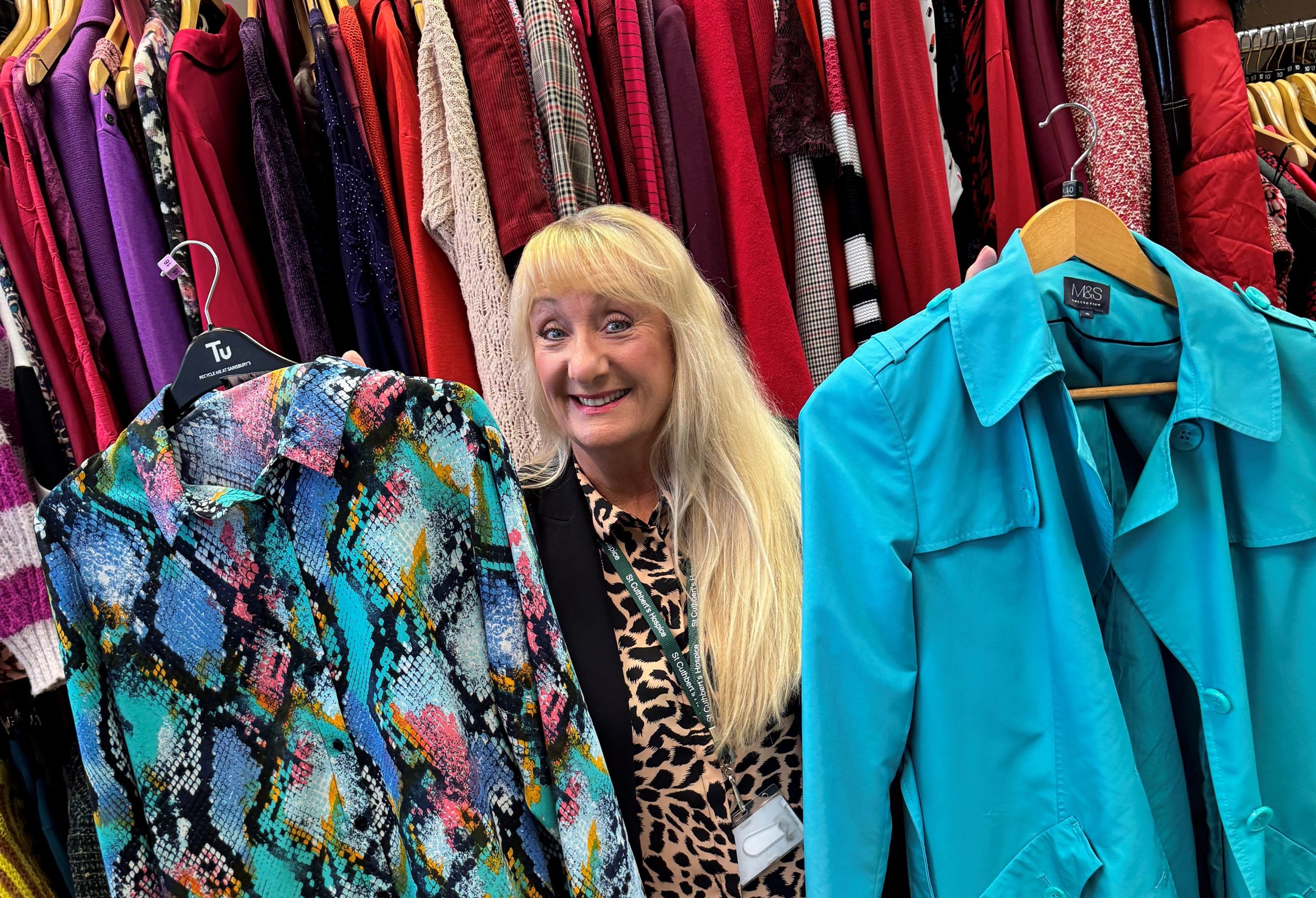 a colourful background of clothes on a rail. a woman stands smiling holding up bright clothes.