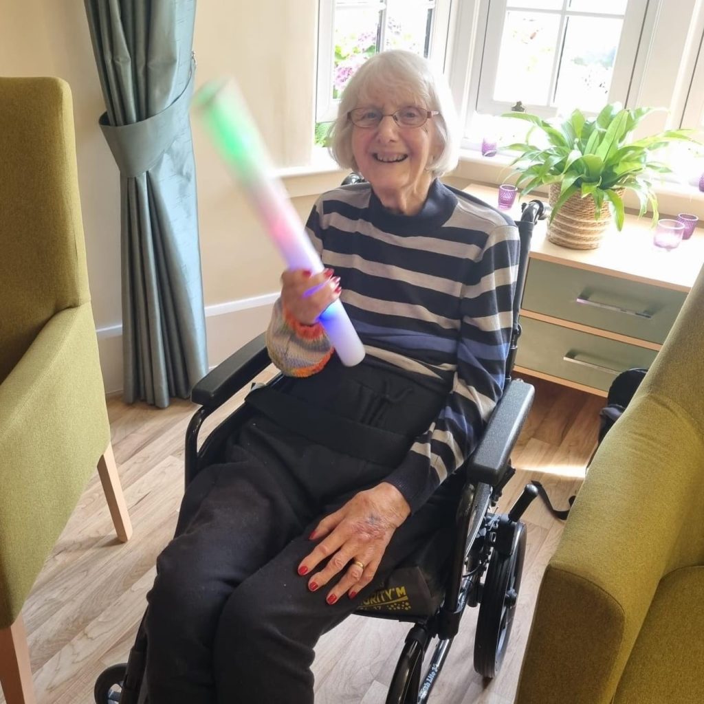 A woman sits in a wheelchair holding a light up toy batton. She smiles broadly.